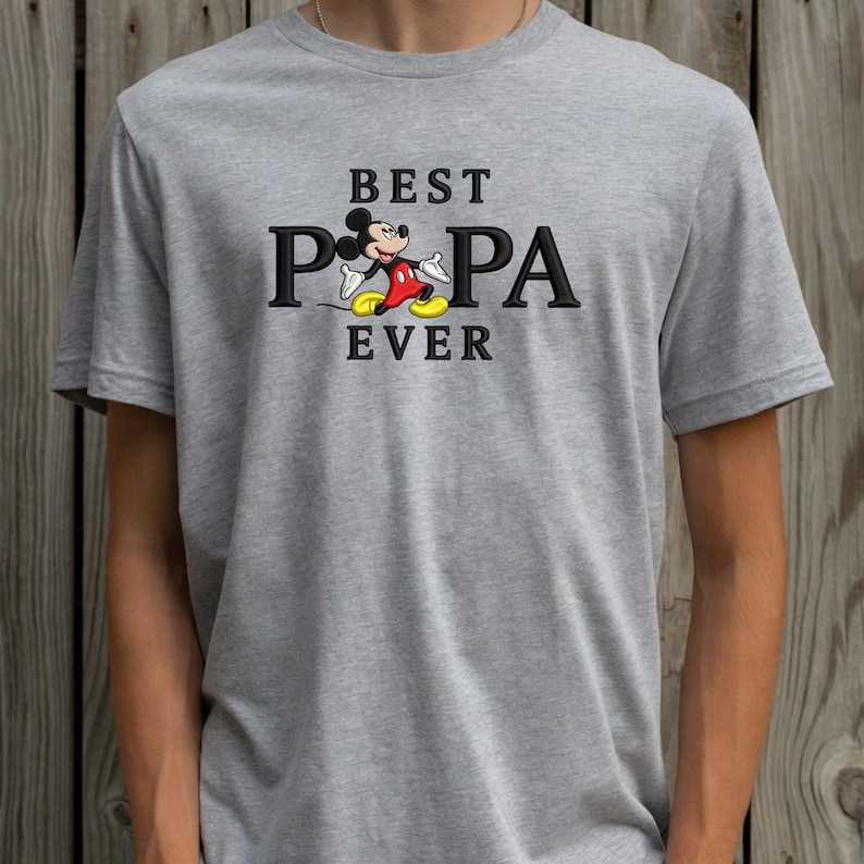 Puede incluir: Camiseta gris con la frase negra "BEST PAPA EVER" y una imagen de Mickey Mouse. Mickey lleva pantalones cortos rojos, zapatos amarillos y guantes blancos. La camiseta es de manga corta y de tejido suave.