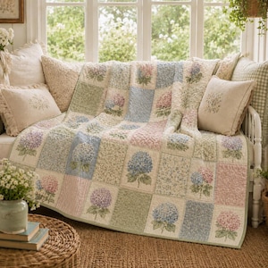 May include: A patchwork quilt featuring a repeating pattern of blue, pink, and purple hydrangea blossoms on a cream background, accented by soft green and floral print squares. The quilt is draped over a white wooden bench with matching throw pillows.