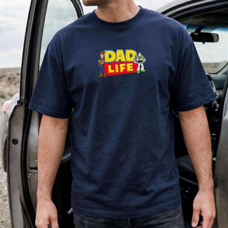 May include: Navy blue t-shirt with the text "DAD LIFE" in yellow and red, featuring cartoon characters. The shirt is short-sleeved and appears to be made of a soft material.