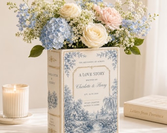 Puede incluir: Un jarrón de cerámica rectangular con forma de libro con ilustraciones de paisajes en azul y blanco. El frente presenta el texto A LOVE STORY, WRITTEN BY Charlotte & Henry, EST. 10.03.2026 y THE BEGINNING OF ALWAYS, lleno de flores en tonos pastel.