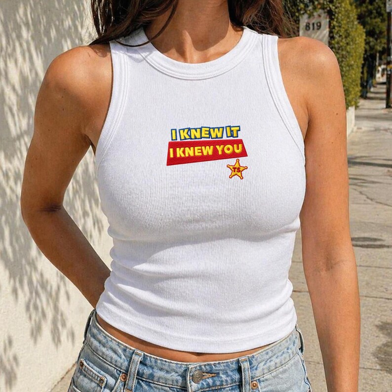 K&ouml;nnte beinhalten: Ein wei&szlig;es, geripptes Tanktop mit einem Grafikdruck auf der Brust. Der Text lautet I KNEW IT in blauen Buchstaben und I KNEW YOU in wei&szlig;en Buchstaben auf rotem Grund. Darunter befindet sich ein kleiner orangefarbener Sheriffstern mit den Buchstaben TS.