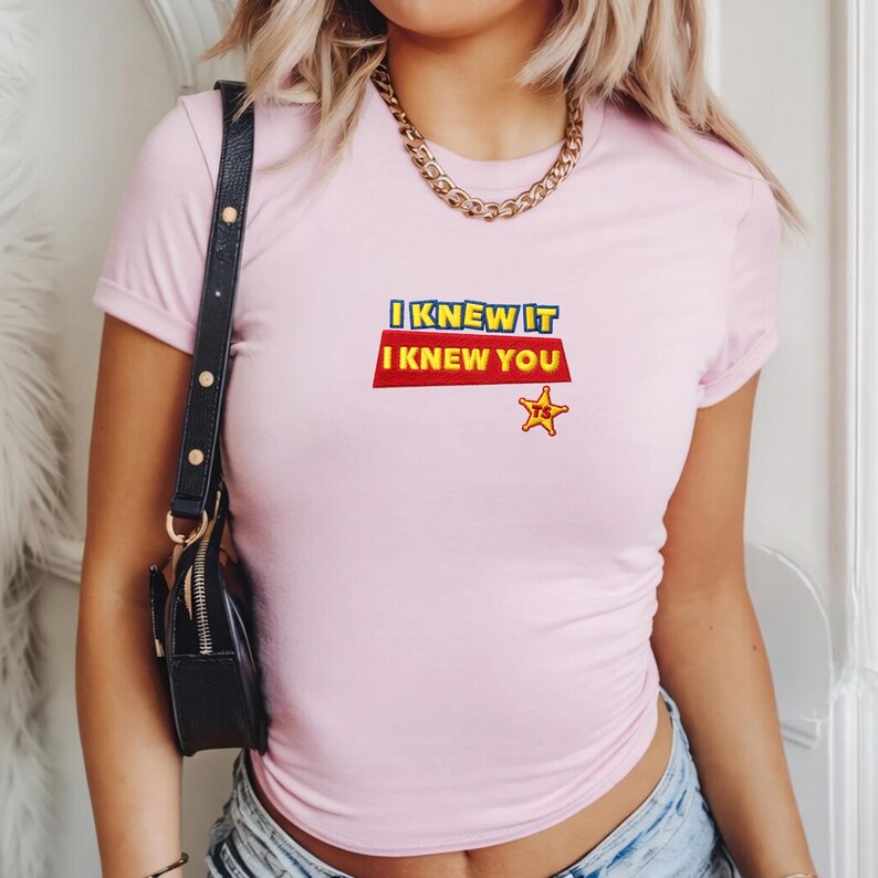 K&ouml;nnte beinhalten: Ein hellrosa T-Shirt mit kurzen &Auml;rmeln, das den Text I KNEW IT I KNEW YOU in Blau und Gelb auf rotem Hintergrund zeigt, begleitet von einem kleinen gelben Sheriff-Stern-Symbol mit den Buchstaben TS in der Mitte des Sterns.