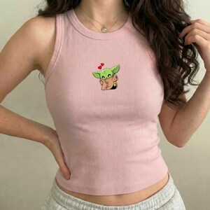 May include: A pale pink ribbed vest top with a cartoon image of a green alien character and heart accents. The sleeveless top has a round neckline and is made of a soft, ribbed material.