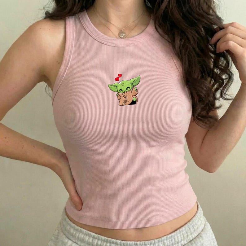 May include: A pale pink ribbed vest top with a cartoon image of a green alien character and heart accents. The sleeveless top has a round neckline and is made of a soft, ribbed material.