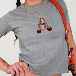 Puede incluir: Una camiseta gris jaspeado con un dibujo animado de una vaquera montada a caballo. La vaquera lleva un sombrero rojo y el caballo es marr&oacute;n con cascos rojos. La camiseta es de estilo casual.