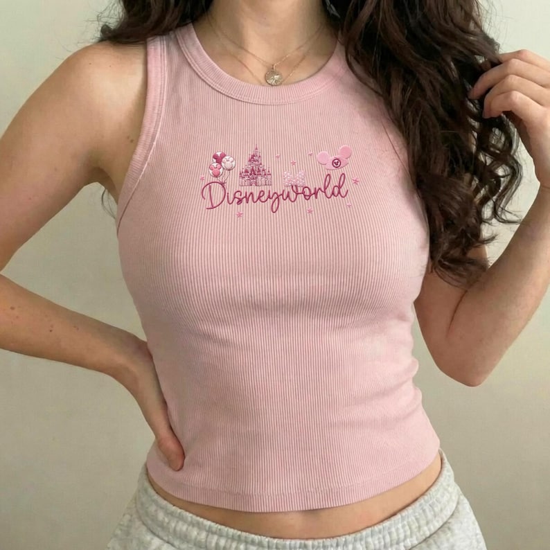 Peut inclure: Un d&eacute;bardeur rose clair c&ocirc;tel&eacute; avec le mot "Disneyworld" en &eacute;criture cursive. Le haut pr&eacute;sente un dessin d'un ch&acirc;teau et de ballons. Le d&eacute;bardeur est sans manches et a une encolure ronde.