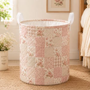 May include: A cylindrical laundry hamper featuring a quilted patchwork design in shades of pink, cream, and beige. The exterior displays alternating squares of floral patterns and plaid textures. It includes two white fabric handles on the sides.