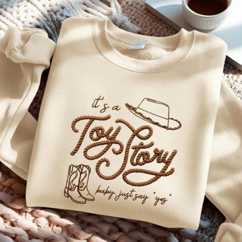 Peut inclure: Un sweat-shirt &agrave; col rond couleur cr&egrave;me avec un motif graphique marron. Le texte "it's a Toy Story" est &eacute;crit dans une police style corde, accompagn&eacute; du contour d'un chapeau de cowboy, de bottes et du texte "baby just say yes".
