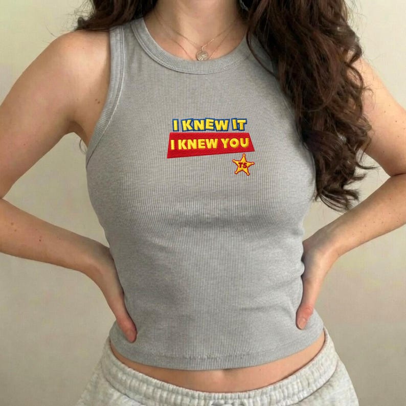Op de afbeelding: Een grijs geribbelde tanktop met de tekst I KNEW IT I KNEW YOU in blauwe en gele letters op een rode rechthoekige achtergrond, vergezeld van een kleine gele stergrafiek met de letters TS in het rood gedrukt in de ster.