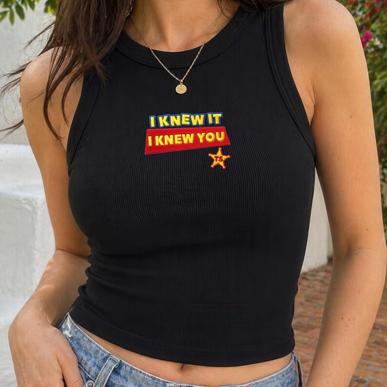 K&ouml;nnte beinhalten: Ein schwarzes geripptes Tanktop mit einem Grafikdruck, der den Text I KNEW IT I KNEW YOU in gelben und wei&szlig;en Buchstaben auf rotem Hintergrund zeigt, erg&auml;nzt durch einen kleinen gelben Sheriffstern mit den Buchstaben TS im Inneren.