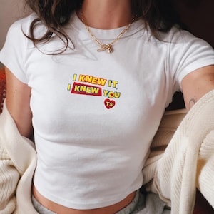 Op de afbeelding: Een wit T-shirt met korte mouwen en geborduurde tekst met de woorden I KNEW IT, I KNEW YOU in gele en blauwe letters op een rode achtergrond, vergezeld van een klein rood hartje met de letters TS, gedragen met een gouden ketting.