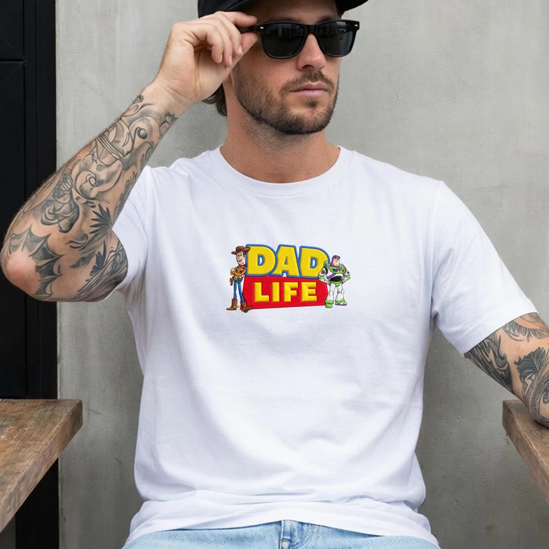 May include: White t-shirt featuring the text "DAD LIFE" in yellow and red, with cartoon characters Woody and Buzz Lightyear. The shirt is a classic crew neck style.