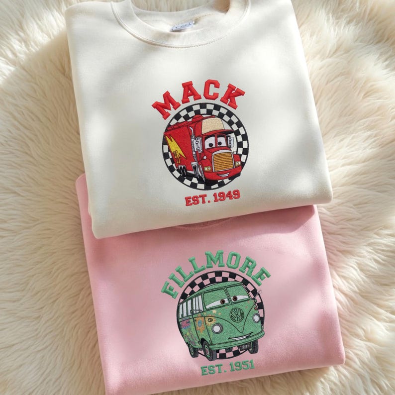 May include: Two crewneck sweatshirts, one cream and one pink, with embroidered cartoon vehicle designs. The cream sweatshirt features a red truck with the word "MACK" and "EST. 1949". The pink sweatshirt has a green van with the word "FILLMORE" and "EST. 1951".