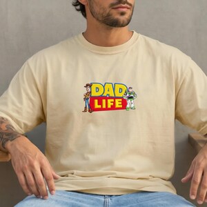 May include: A beige t-shirt featuring the text "DAD LIFE" in yellow and red, with cartoon images of Woody and Buzz Lightyear. The shirt is a casual, short-sleeved garment.