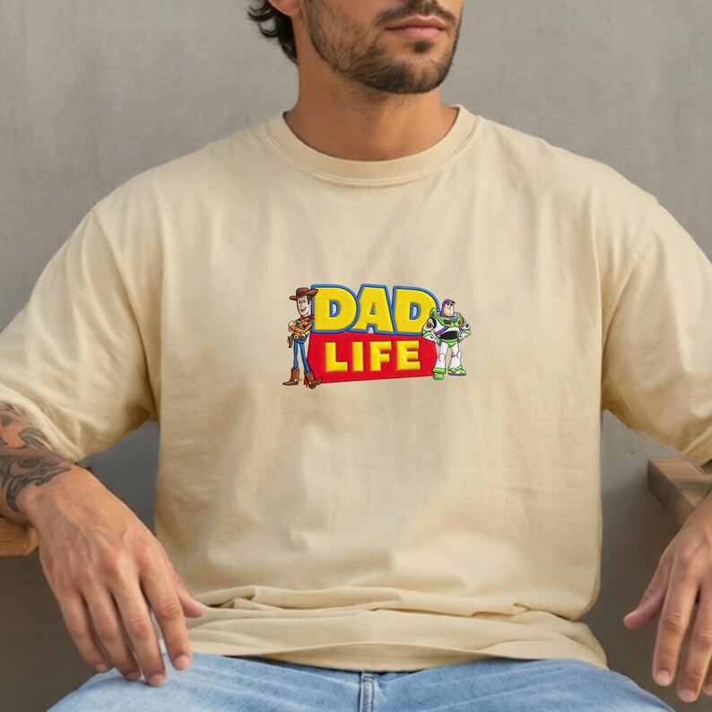 May include: A beige t-shirt featuring the text "DAD LIFE" in yellow and red, with cartoon images of Woody and Buzz Lightyear. The shirt is a casual, short-sleeved garment.