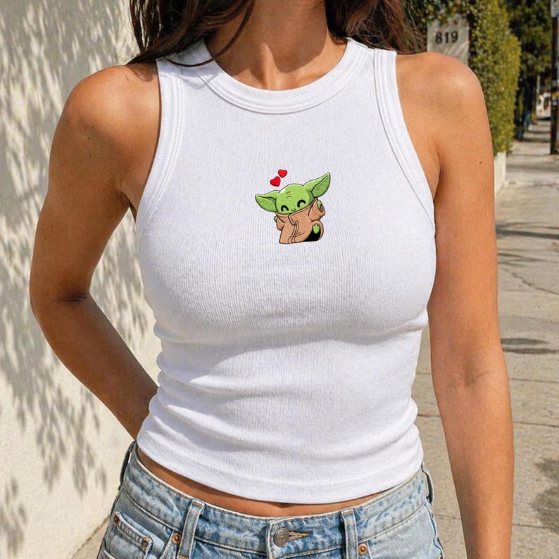May include: White ribbed vest top with a cartoon image of a green alien character holding a brown blanket, with two red hearts above it. The vest top has a round neckline and a sleeveless design.