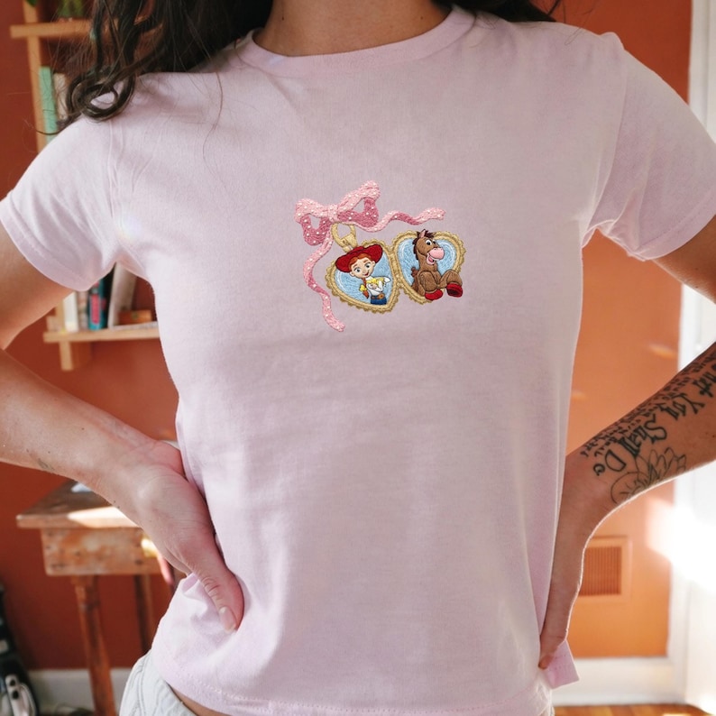 May include: A light pink t-shirt featuring embroidered heart-shaped pendants with cartoon characters. The pendants are connected by a pink bow with white polka dots. The shirt is made of a soft material.