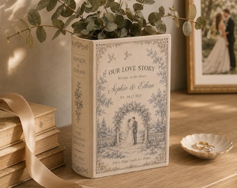 May include: A decorative book-shaped box featuring a cream cover with blue floral illustrations, the text "OUR LOVE STORY", "Sophia & Ethan", "Est. 04.12.2025", and "Every Page Leads Us Home". The spine displays "SOPHIA & ETHAN" and "THE WEDDING EDITION".
