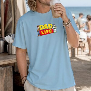 May include: Light blue t-shirt with the text "DAD LIFE" in yellow and red, featuring cartoon characters. The shirt is a casual, short-sleeved style.