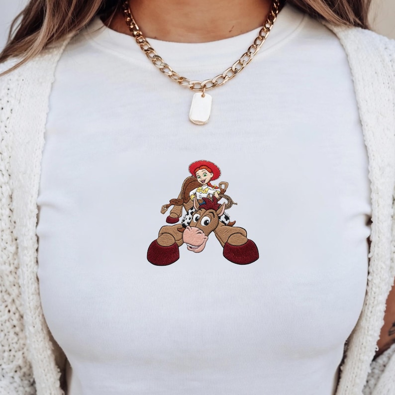 Puede incluir: Camiseta blanca con un dibujo animado de una vaquera montando a caballo. La vaquera lleva un sombrero rojo y una camisa blanca. El caballo es marr&oacute;n con manchas blancas y negras. La camiseta tiene cuello redondo.