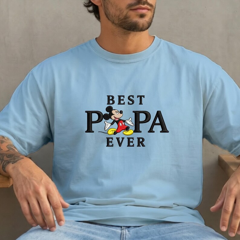 Puede incluir: Camiseta azul claro con el texto "BEST PA PA EVER" en letras negras. La "P" de "PA" se reemplaza con una imagen de dibujos animados de Mickey Mouse con pantalones cortos rojos, zapatos amarillos y guantes blancos.