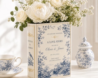 Puede incluir: Un florero rectangular con forma de libro, fondo crema y patrón floral azul estilo chinoiserie. El frente presenta el texto "OUR GREATEST ADVENTURE A LOVE STORY WRITTEN BY OLIVIA & JAMES EST. 09.12.2026 HOME IS WHERE OUR STORY CONTINUES."