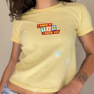May include: A pale yellow short-sleeved t-shirt featuring a central graphic design. The graphic displays three colorful square blocks with the letters T, S, and O, surrounded by the text I KNEW IT and I KNEW YOU printed in a bold red font.