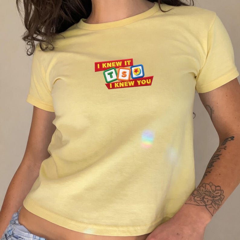 May include: A pale yellow short-sleeved t-shirt featuring a central graphic design. The graphic displays three colorful square blocks with the letters T, S, and O, surrounded by the text I KNEW IT and I KNEW YOU printed in a bold red font.