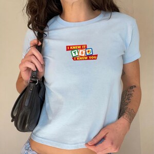 May include: A light blue short-sleeved t-shirt featuring a graphic print on the chest. The design includes the text "I KNEW IT" and "I KNEW YOU" in red banners, surrounding three colorful square blocks displaying the letters T, S, and a ball icon.