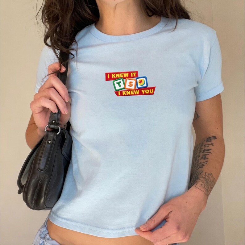 May include: A light blue short-sleeved t-shirt featuring a graphic print on the chest. The design includes the text "I KNEW IT" and "I KNEW YOU" in red banners, surrounding three colorful square blocks displaying the letters T, S, and a ball icon.
