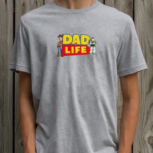 May include: A gray t-shirt featuring the text "DAD LIFE" in yellow and red, with cartoon illustrations of Woody and Buzz Lightyear. The shirt has short sleeves and a crew neck.