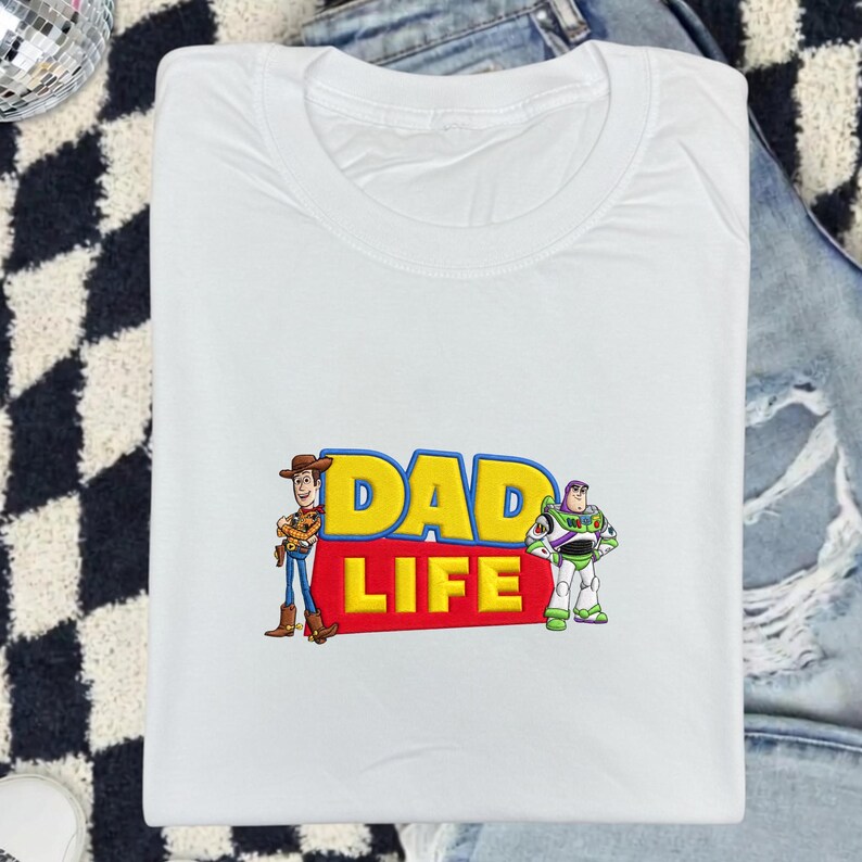 May include: White t-shirt with the text "DAD LIFE" in yellow and red, featuring cartoon characters Woody and Buzz Lightyear. The shirt is folded on a patterned surface with a pair of jeans.