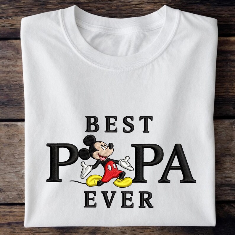 Puede incluir: Camiseta blanca con el texto negro "BEST PA EVER", con el personaje de dibujos animados Mickey Mouse. Mickey lleva pantalones cortos rojos, zapatos amarillos y guantes blancos. La camiseta est&aacute; doblada sobre una superficie de madera.