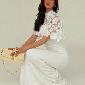 May include: A woman in a white dress and lace top, holding a cream-colored quilted handbag with a gold chain strap. A gold flower ring adorns the hand. The dress is floor-length.