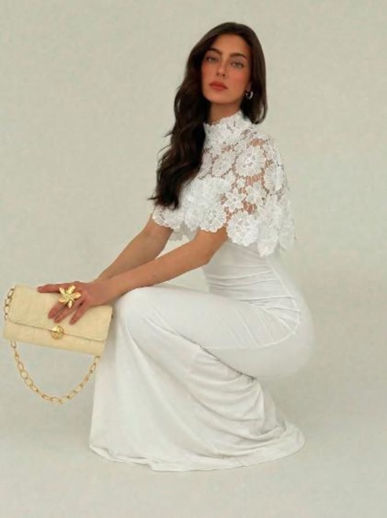 May include: A woman in a white dress and lace top, holding a cream-colored quilted handbag with a gold chain strap. A gold flower ring adorns the hand. The dress is floor-length.