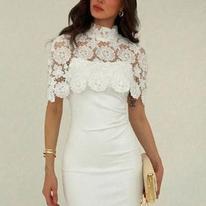 May include: A white, form-fitting dress with a high neckline. The dress features a delicate, white lace overlay on the upper bodice and sleeves. A gold clutch and bracelet complete the look.