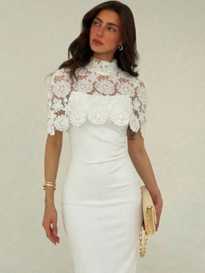 May include: A white, form-fitting dress with a high neckline. The dress features a delicate, white lace overlay on the upper bodice and sleeves. A gold clutch and bracelet complete the look.