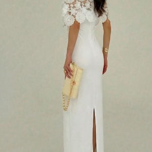 May include: A white, floor-length gown with a high neckline and a slit up the back. The dress is paired with a cream-colored clutch with a gold chain strap and a white lace capelet.