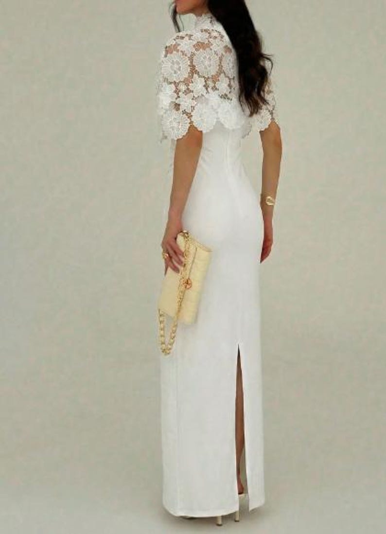 May include: A white, floor-length gown with a high neckline and a slit up the back. The dress is paired with a cream-colored clutch with a gold chain strap and a white lace capelet.