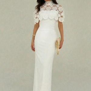 May include: A white, floor-length gown with a high neckline and a lace overlay on the bodice. The dress features a fitted silhouette and is paired with a gold clutch. The gown is suitable for formal events.