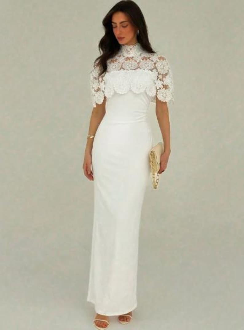 May include: A white, floor-length gown with a high neckline and a lace overlay on the bodice. The dress features a fitted silhouette and is paired with a gold clutch. The gown is suitable for formal events.
