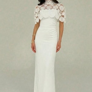 May include: A white, floor-length gown with a high neckline and a delicate lace overlay. The dress features a fitted silhouette and a subtle ruched detail at the waist. The lace capelet adds a touch of elegance.