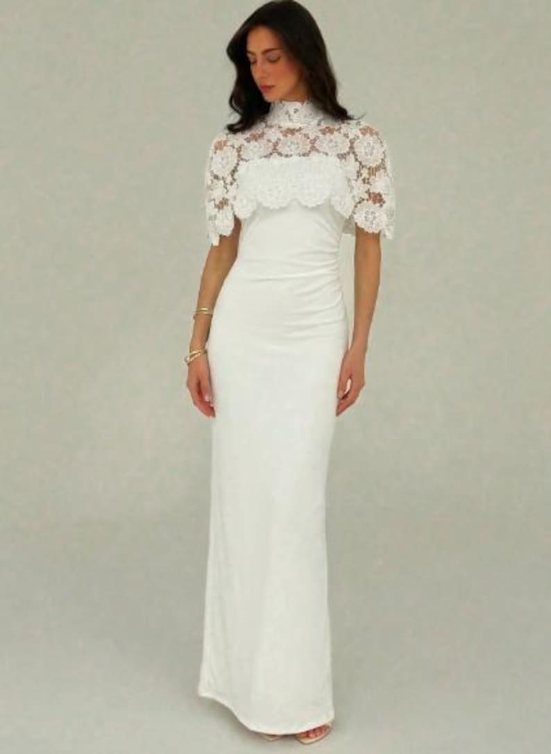 May include: A white, floor-length gown with a high neckline and a delicate lace overlay. The dress features a fitted silhouette and a subtle ruched detail at the waist. The lace capelet adds a touch of elegance.