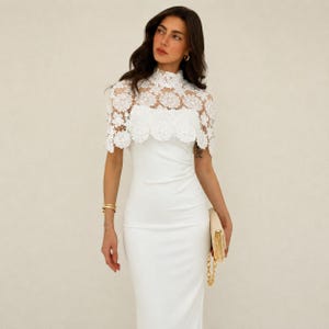 May include: A white, floor-length dress with a high neckline and a delicate, lace overlay. The lace detail extends to the shoulders, creating a cape-like effect. A gold clutch with a chain strap is held in the hand.