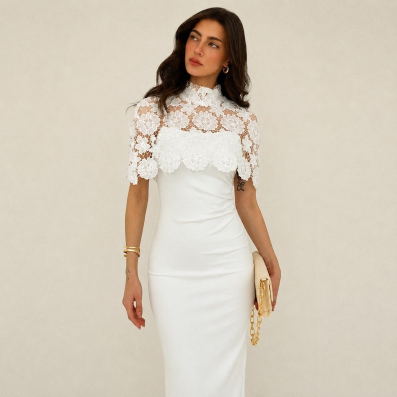 May include: A white, floor-length dress with a high neckline and a delicate, lace overlay. The lace detail extends to the shoulders, creating a cape-like effect. A gold clutch with a chain strap is held in the hand.