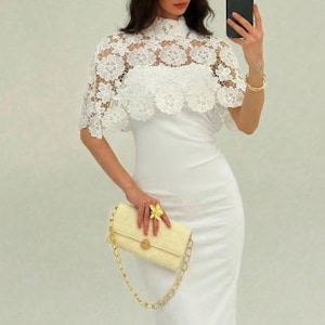 May include: A white, form-fitting dress with a high neckline, paired with a white lace capelet. A light yellow handbag with a gold chain strap and a gold flower ring complete the look. The outfit is elegant and suitable for special occasions.