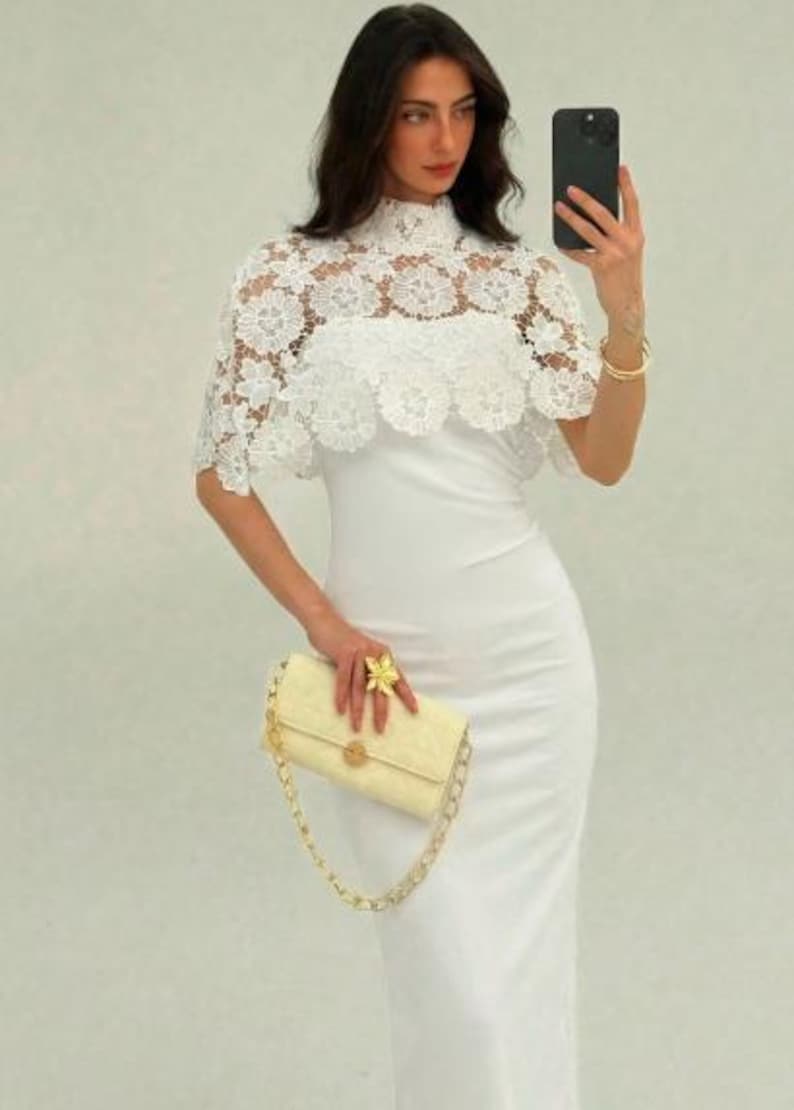 May include: A white, form-fitting dress with a high neckline, paired with a white lace capelet. A light yellow handbag with a gold chain strap and a gold flower ring complete the look. The outfit is elegant and suitable for special occasions.