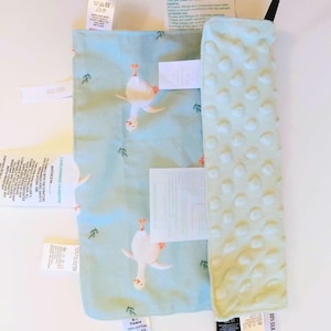 May include: A rectangular baby security blanket featuring a light blue fabric with a repeating white goose pattern and a contrasting pale green textured minky side with raised dots. Multiple white care and safety instruction labels are attached.