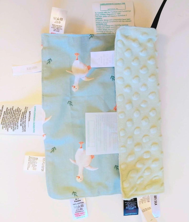 May include: A rectangular baby security blanket featuring a light blue fabric with a repeating white goose pattern and a contrasting pale green textured minky side with raised dots. Multiple white care and safety instruction labels are attached.