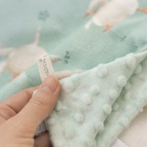 May include: A light mint green baby blanket featuring a repeating pattern of white geese. The fabric has a textured dimpled surface on one side and a smooth side with the printed geese. A small white fabric tag with black text is attached to the edge.